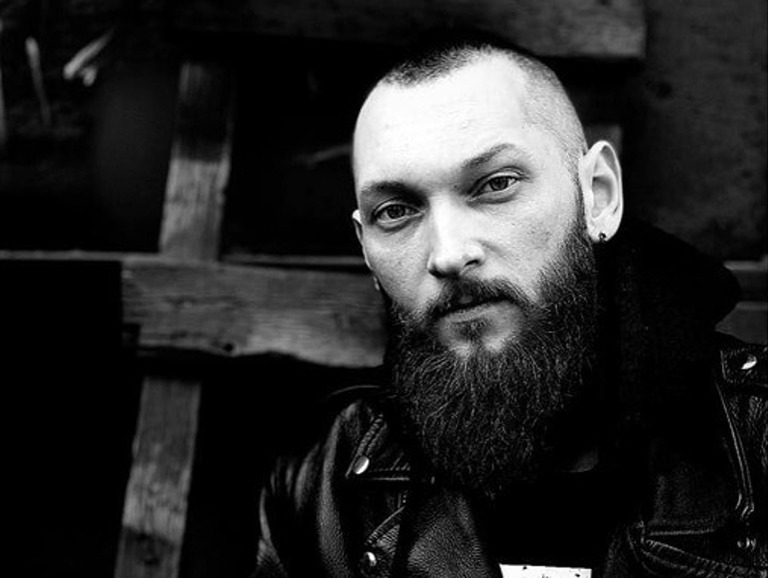 Schwarz-Weiß-Porträt eines Mannes mit Glatze, Bart und Lederjacke, der direkt in die Kamera blickt.