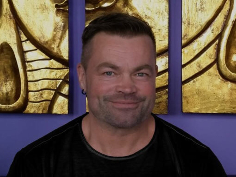 Ein lächelnder Mann mit kurzem Haar und Bartstoppeln trägt ein schwarzes Hemd vor goldener Buddha-Wandkunst.